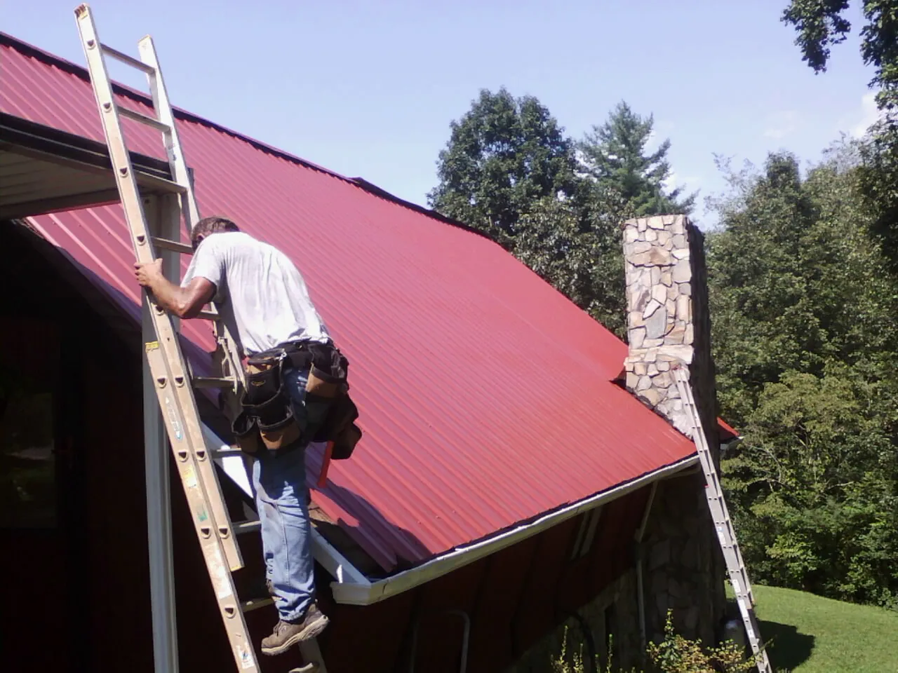 Roofer on a ladder beside a red metal roof and stone chimney — Soffit & Fascia Repair by Roofing Brothers in Lockwood